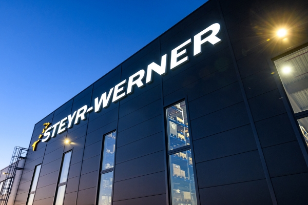 Vor dem klaren Abendhimmel erstrahlt die moderne Fassade von „STEYR-WERNER“. Große, vertikale Fenster setzen den Innenraum in Szene, während von innen ein sanftes, warmes Licht hereinströmt.